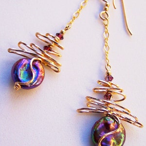 May include: Gold-coloured drop earrings with iridescent, purple coin-shaped beads. The earrings have a spiral wire design and are accented with small, purple beads. The ear wires and chains are also gold-coloured.