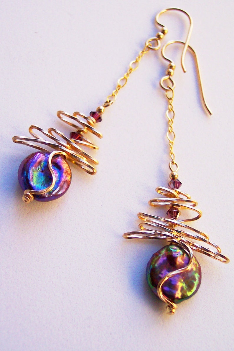 May include: Gold-coloured drop earrings with iridescent, purple coin-shaped beads. The earrings have a spiral wire design and are accented with small, purple beads. The ear wires and chains are also gold-coloured.