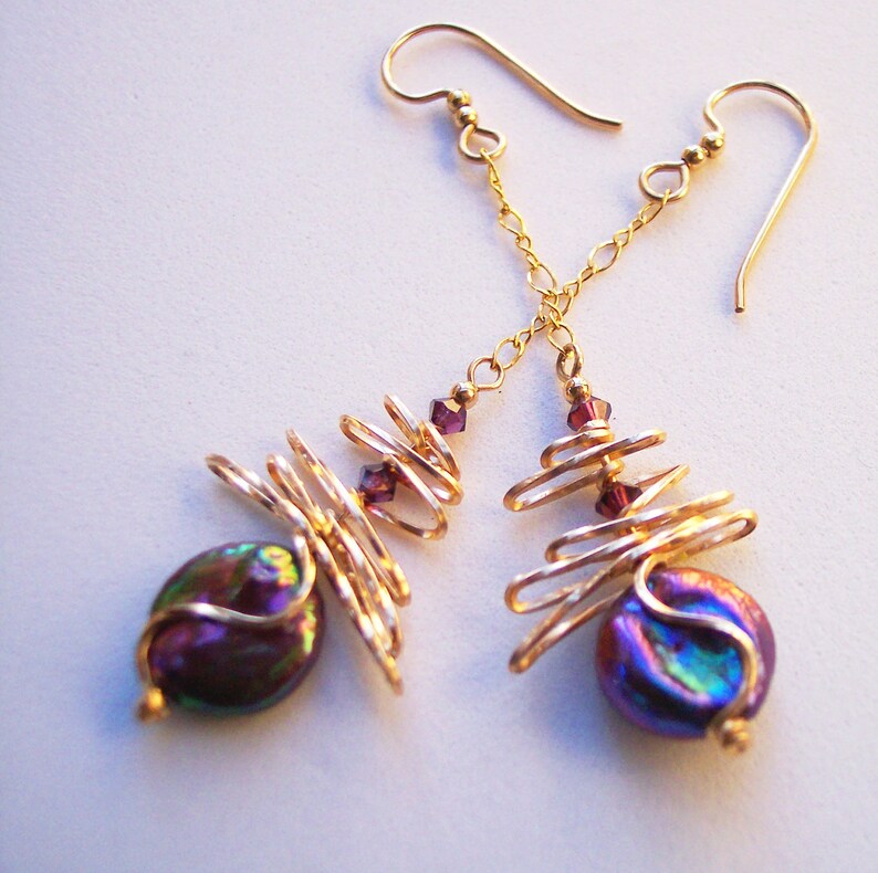 May include: Gold-coloured earrings with iridescent, dark purple, oval beads. The earrings have a spiral wire design with small, purple crystal accents and gold-coloured chain details.