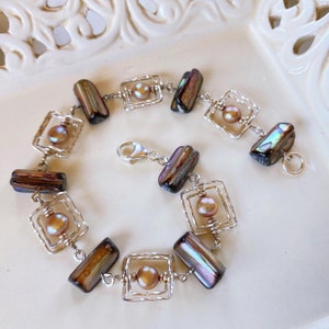 Op de afbeelding: Een zilveren armband met iriserende, rechthoekige kralen en ronde, perzik-kleurige parels. De kralen zijn verbonden door zilverdraad, met elke parel gezet in een vierkant frame. De armband heeft een karabijnsluiting.