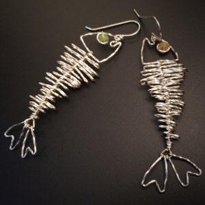 May include: A pair of silver-toned wire earrings in the shape of fish skeletons. Each earring features a coiled body, a head with a gemstone, and a tail fin. The earrings are displayed against a dark background.