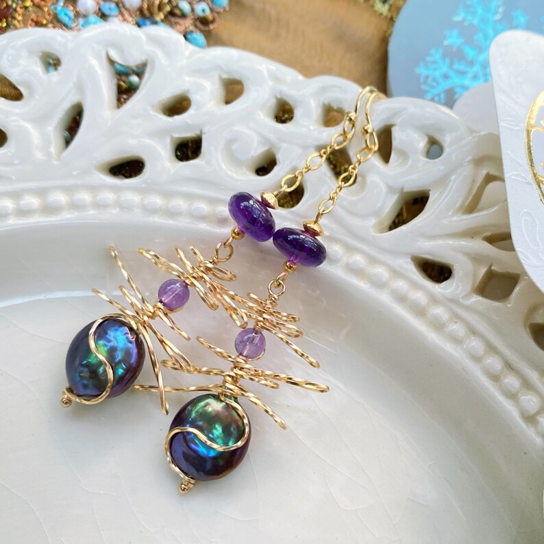 May include: Dangle earrings in gold, with iridescent dark pearls and purple amethyst beads. The earrings have a unique wire design and are suspended from delicate gold chains, showcasing a blend of natural elements and artistic craftsmanship.
