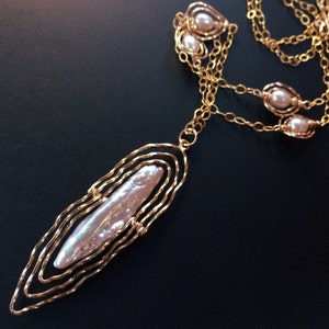 May include: A gold necklace featuring a large, elongated pearl pendant. The pendant is set within a layered, wavy gold frame. Smaller pearls are also set along the chain. The necklace is shown against a dark background.