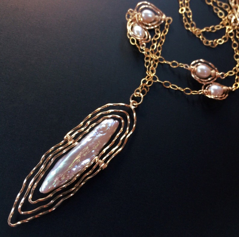 May include: A gold necklace featuring a large, elongated pearl pendant. The pendant is set within a layered, wavy gold frame. Smaller pearls are also set along the chain. The necklace is shown against a dark background.