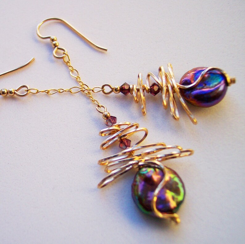 May include: Gold earrings with a spiral design and iridescent, dark purple-blue beads. The earrings feature small, round, purple crystals and delicate chain accents. The design is abstract and modern, with a focus on the interplay of light and colour.