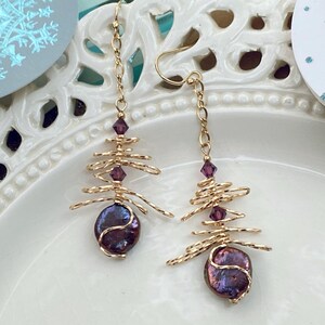 May include: A pair of gold-coloured wire earrings with a distinctive design. Each earring has a dark purple, iridescent pearl at the base, a wire tree-like structure, and purple crystal accents. The earrings are suspended from gold-coloured ear wires.