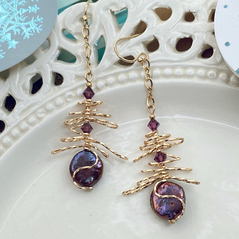 May include: A pair of gold-coloured wire earrings with a distinctive design. Each earring has a dark purple, iridescent pearl at the base, a wire tree-like structure, and purple crystal accents. The earrings are suspended from gold-coloured ear wires.