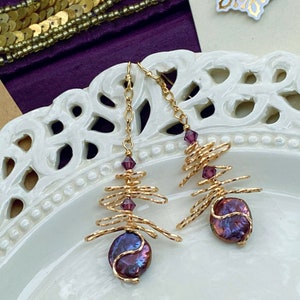 May include: A pair of gold-coloured wire earrings. Each earring has a spiral wire design, small purple crystals, and a large, iridescent, purple-blue pearl. The earrings are suspended from fine gold chains.