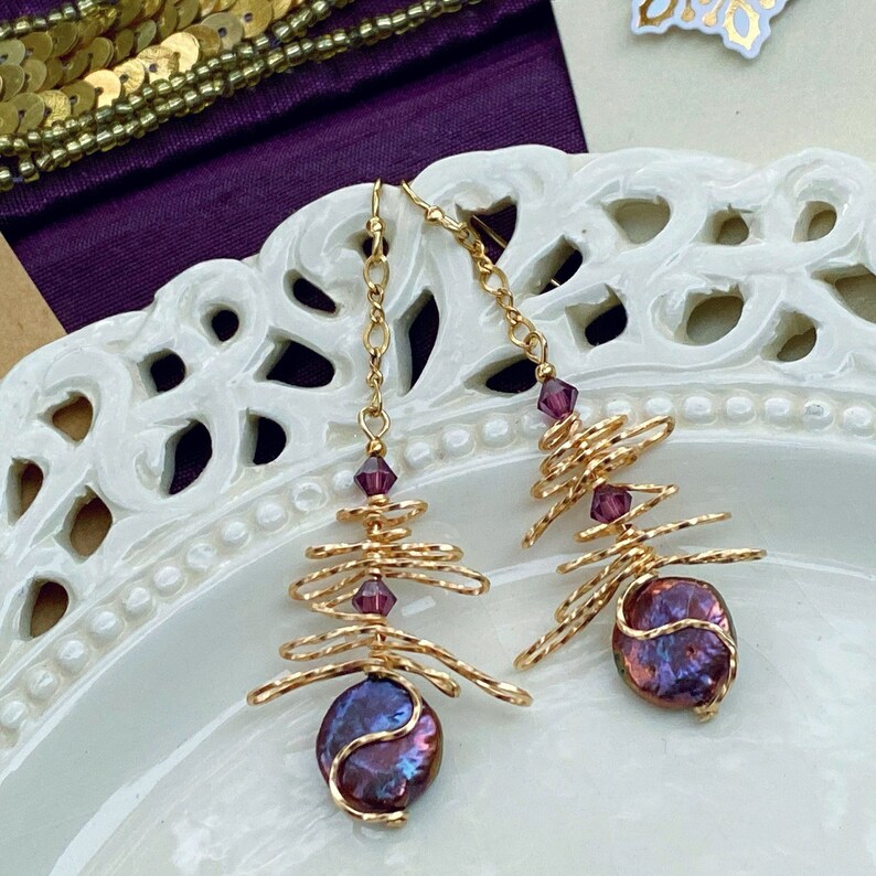May include: A pair of gold-coloured wire earrings. Each earring has a spiral wire design, small purple crystals, and a large, iridescent, purple-blue pearl. The earrings are suspended from fine gold chains.