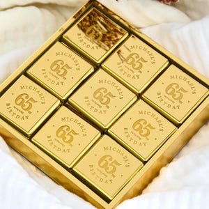 May include: A gold-colored box filled with square chocolates. Each chocolate is engraved with "MICHAEL'S BIRTHDAY 65" and the date. The chocolates are arranged in a grid pattern, perfect for a milestone birthday celebration.