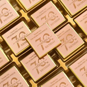 May include: Close-up of square, pink chocolates with gold trim, arranged in a gold tray. Each chocolate is engraved with "Thank You for Celebrating with Us" and "Nelson's 70th Birthday". A celebratory gift.