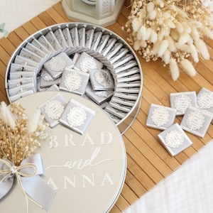 May include: A round, silver-colored container filled with individually wrapped chocolates. The chocolates are adorned with a decorative label that reads "Brad and Anna." The container sits on a wooden surface, with additional chocolates scattered nearby. A bouquet of dried flowers and a decorative lantern are also present.