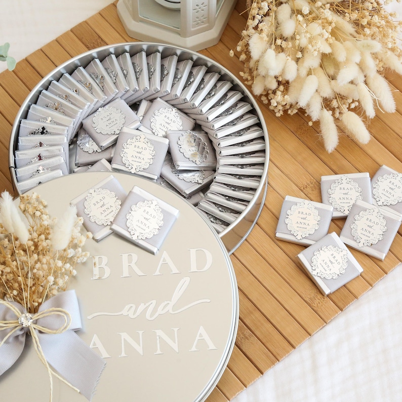 May include: A round, silver-colored container filled with individually wrapped chocolates. The chocolates are adorned with a decorative label that reads "Brad and Anna." The container sits on a wooden surface, with additional chocolates scattered nearby. A bouquet of dried flowers and a decorative lantern are also present.
