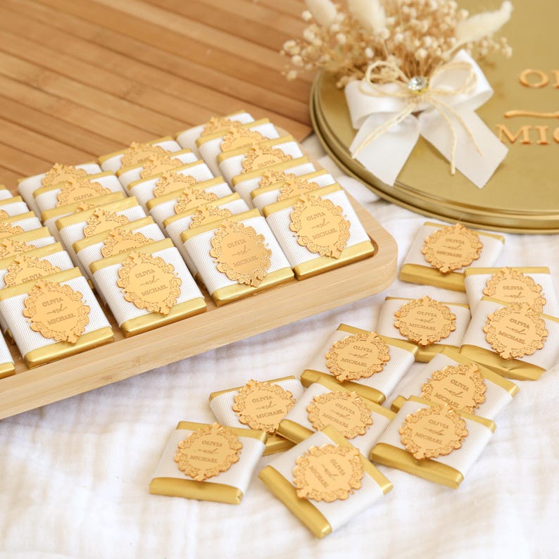May include: A collection of chocolates, each wrapped in white and gold foil, arranged on a wooden tray. Each chocolate is adorned with a gold-colored decorative label. A gold-colored round container with a white bow and dried flowers is in the background.