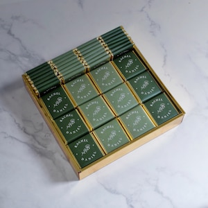 Op de afbeelding: Een gouden doos gevuld met kleine groene en gouden chocolade reepjes. Elke reep is verpakt met de namen "Rachel" en "Daniel" erop gedrukt.