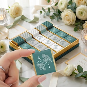 Op de afbeelding: Een teal en gouden doosje chocolade met een bloemenontwerp. De chocolaatjes zijn teal en wit met een bloemenontwerp. Eén chocolaatje wordt omhoog gehouden, met de namen "Anna Jacob" en de letters "A" en "J".
