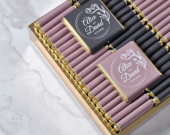Può includere: Una scatola di barrette di cioccolato avvolte singolarmente con un bordo dorato. Le barrette di cioccolato sono avvolte in una lamina rosa chiaro con un bordo dorato. Le due barrette superiori hanno un involucro nero con un design bianco che dice "Alice e David 11.11.2024".