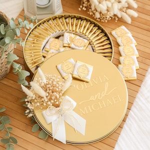 K&ouml;nnte beinhalten: Eine goldfarbene runde Geschenkbox gef&uuml;llt mit einzeln verpackten Pralinen. Der Deckel ist mit "Olivia and Michael" graviert und mit einer wei&szlig;en Schleife und Trockenblumen verziert. Die Pralinen haben eine goldene und wei&szlig;e Verpackung.