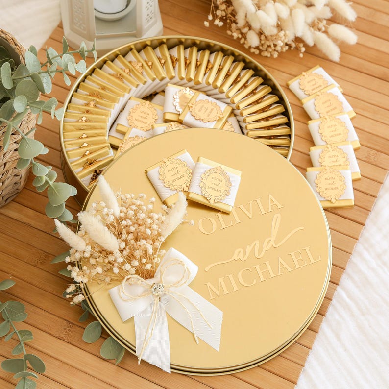 K&ouml;nnte beinhalten: Eine goldfarbene runde Geschenkbox gef&uuml;llt mit einzeln verpackten Pralinen. Der Deckel ist mit "Olivia and Michael" graviert und mit einer wei&szlig;en Schleife und Trockenblumen verziert. Die Pralinen haben eine goldene und wei&szlig;e Verpackung.