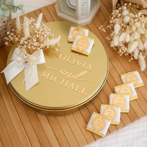 May include: A gold-colored round tin with the names "OLIVIA and MICHAEL" embossed on the lid. The tin is decorated with a white ribbon, dried flowers, and small chocolates. The chocolates have gold labels.