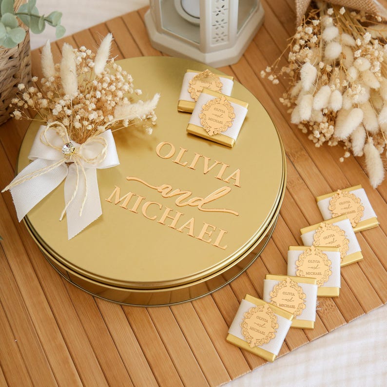 May include: A gold-colored round tin with the names "OLIVIA and MICHAEL" embossed on the lid. The tin is decorated with a white ribbon, dried flowers, and small chocolates. The chocolates have gold labels.