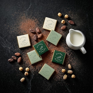 May include: An overhead shot of gourmet chocolates, cocoa powder, nuts, and a small pitcher of milk on a dark surface. The chocolates are in green and cream-colored wrappers, each with the text "RACHEL DANIEL". Cocoa beans and nuts are scattered around the chocolates.