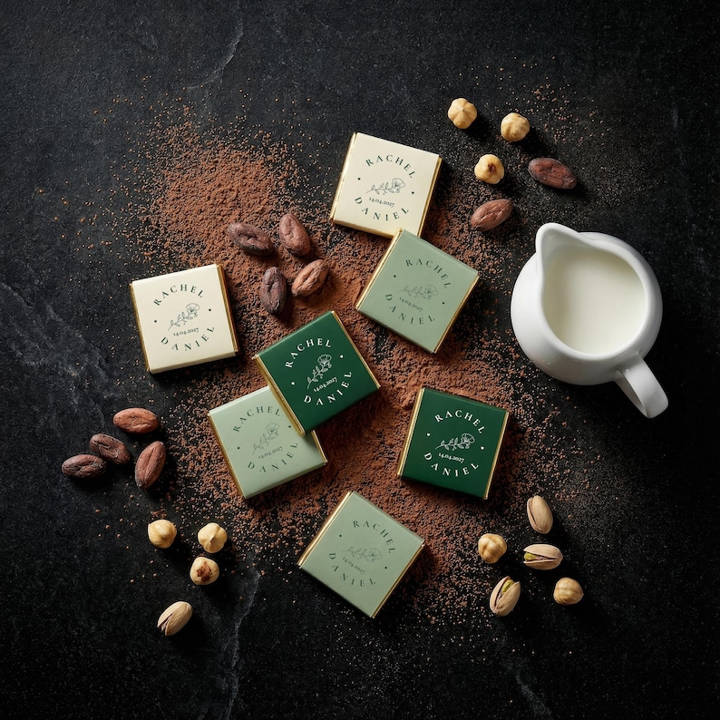 May include: An overhead shot of gourmet chocolates, cocoa powder, nuts, and a small pitcher of milk on a dark surface. The chocolates are in green and cream-colored wrappers, each with the text "RACHEL DANIEL". Cocoa beans and nuts are scattered around the chocolates.