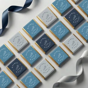 May include: An overhead shot of several square chocolates with gold foil edges. Each chocolate has a different shade of blue or gray paper with the text "RACHEL DANIEL" and a floral design. Ribbons in blue and gray are in the background.