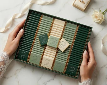 May include: A dark green rectangular box filled with chocolate bars in varying shades of green and cream. The box is decorated with gold trim and the text "RACHEL DANIEL". A small ring box and a white rose are also present.