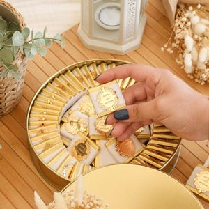 May include: A round, gold-colored tin filled with individually wrapped chocolates. Each chocolate is square, with a white wrapper and gold foil accents. The chocolates are arranged in a circular pattern. The top of the tin is also gold. The chocolates have the text "Olivia and Michael".