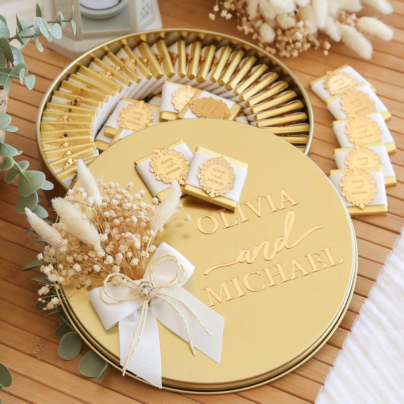May include: A gold-colored round box with the words "OLIVIA and MICHAEL" engraved on the lid. The box is filled with chocolates, some wrapped in white and gold paper. A small bouquet of dried flowers and a white ribbon adorn the box.