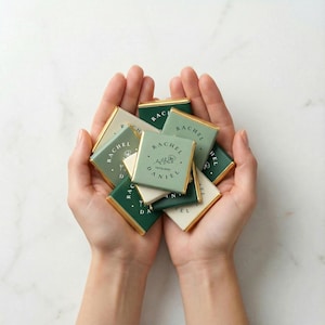 May include: A handful of small, square chocolates with gold foil edges. The chocolates are in shades of green and white, with the name "RACHEL DANIEL" and a floral design printed on them. The chocolates are a potential wedding favor.