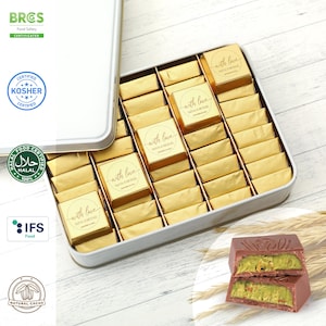 May include: A silver tin box filled with individually wrapped gold foil chocolates. The chocolates are labelled with the text "with love" and the names Olivia & Michael. The box is sitting on a wooden surface with wheat stalks in the foreground. A close-up of a broken chocolate bar with pistachio filling is also visible.
