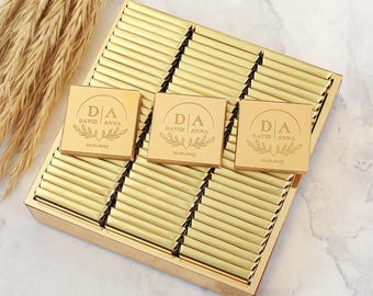 Könnte beinhalten: Eine goldene Schachtel gefüllt mit einzeln verpackten Schokoladentafeln. Jede Tafel ist mit einer goldenen Folie umhüllt und mit einem personalisierten Etikett mit den Initialen "D.A." und den Namen "David & Anna" sowie dem Datum "10.20.2025" versehen.