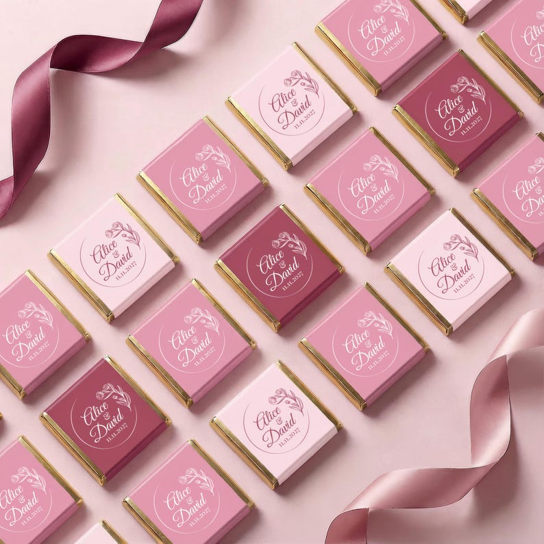 May include: Arrangement of pink and burgundy chocolate bars with gold foil edges. Each bar features the names "Alice & David" and the date "11.11.2027" in a decorative font. A pink ribbon is draped across the scene.