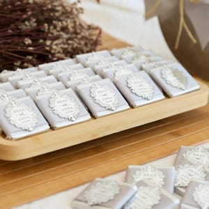 May include: A wooden tray holds numerous small, wrapped chocolates. Each chocolate is wrapped in light gray paper and adorned with a decorative white label that reads "Brad and Anna". The chocolates are arranged neatly, suggesting a wedding or event favor.