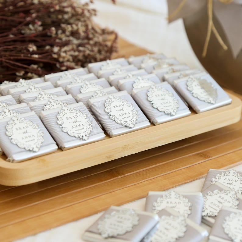 May include: A wooden tray holds numerous small, wrapped chocolates. Each chocolate is wrapped in light gray paper and adorned with a decorative white label that reads "Brad and Anna". The chocolates are arranged neatly, suggesting a wedding or event favor.