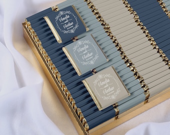 May include: A box of individually wrapped chocolate bars with a gold trim. The wrappers are a combination of light blue and beige with a white floral design and the text "Amelia and Arthur 10.10.2024".