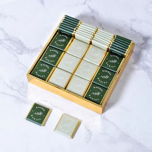Op de afbeelding: Een gouden doos gevuld met groene en witte chocoladevierkanten. Elk vierkant is versierd met de namen "Rachel" en "Daniel" in witte letters. De doos staat op een wit marmeren oppervlak.