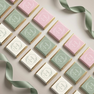 May include: An overhead shot displays an array of personalized chocolate bars. The bars are in pastel pink, sage green, and white, with gold foil edges. Each bar features the text "EMMA & LUCAS" within a geometric frame, with the year 2023.