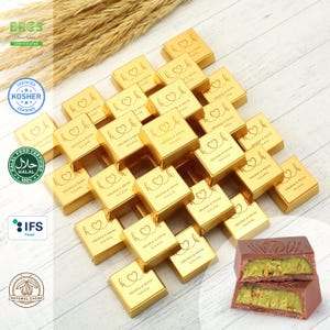 May include: A large group of gold-colored chocolate boxes with the text "HELENA & KEVIN" and a date printed on each box. The boxes are arranged in a pile on a wooden surface. Two pieces of chocolate with pistachio filling are visible in the bottom right corner of the image.