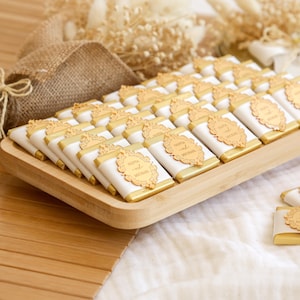 K&ouml;nnte beinhalten: Ein Holztablett gef&uuml;llt mit einzeln verpackten Pralinen. Jede Praline ist in wei&szlig;er und goldener Folie verpackt und mit einem goldenen Etikett mit der Aufschrift "Olivia and Michael" versehen. Die Pralinen sind ordentlich angeordnet, passend f&uuml;r eine Feier.