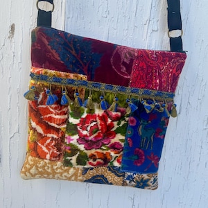 May include: A crossbody bag featuring a patchwork design of velvet and fabric panels in shades of red, blue, green, and gold. The bag includes a decorative blue trim with small hanging tassels and a black adjustable strap with metal rings.