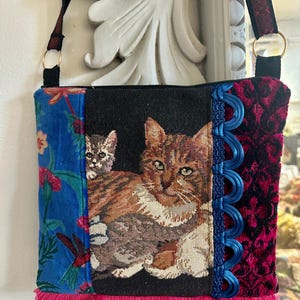 May include: A tapestry shoulder bag featuring a cat design. The bag has a black background with an image of a cat and kitten. The bag has a blue floral pattern on one side and a red velvet pattern on the other. The bag has a pink fringe.