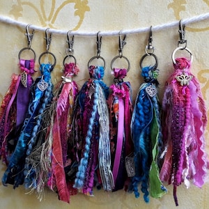 May include: Seven colorful fabric tassel keychains with metal rings and charms. The tassels are made of various shades of pink, purple, blue, and green fabric.