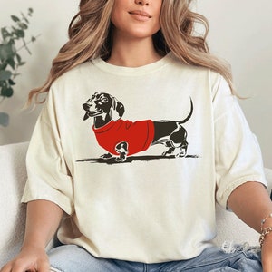 A woman with long wavy balayage hair sits casually on a chair in a modern living room. She is wearing boyfriend jeans with rips in the knees and an oversized Ivory t-shirt with artwork of a smiling dachshund in a red sweater on it.