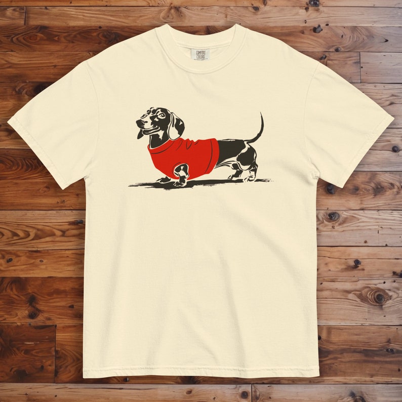 May include: A white t-shirt featuring a black and white illustration of a dachshund wearing a red jumper.