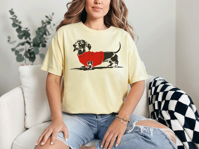 May include: A yellow t-shirt featuring a black and white illustration of a dachshund wearing a red sweater.