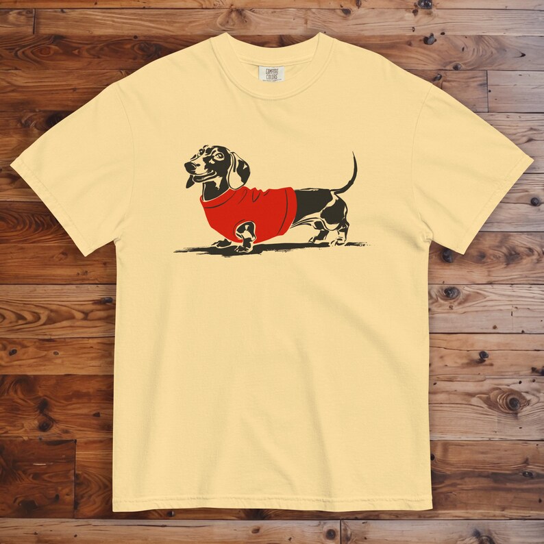 May include: A yellow t-shirt featuring a black and white illustration of a dachshund wearing a red sweater.