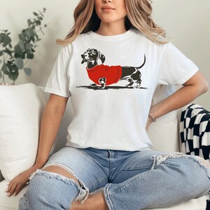 May include: A white t-shirt featuring a black and white illustration of a dachshund wearing a red jumper.
